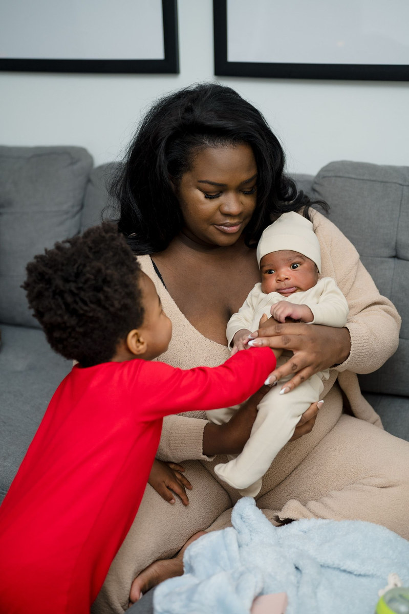 Woman holding a newborn as a young boy in red reaches for the baby