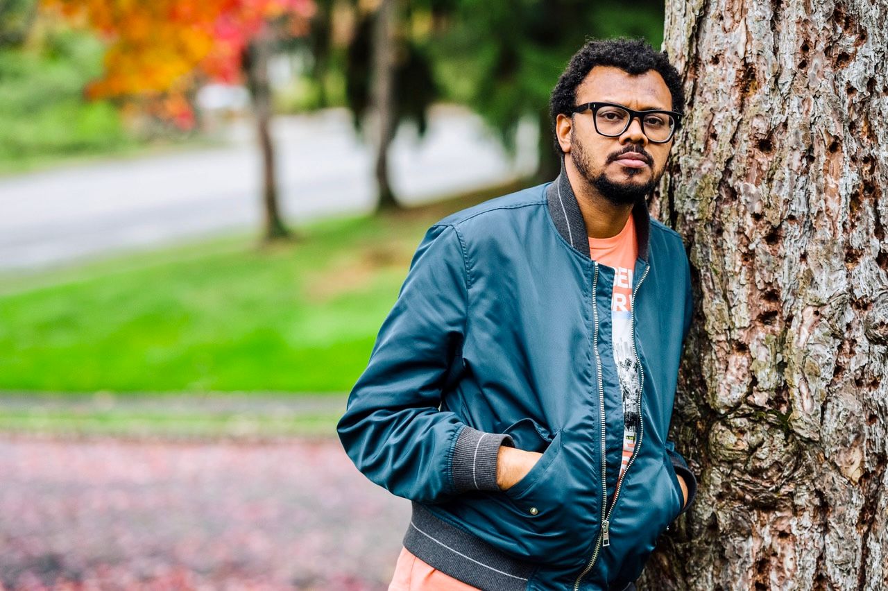 Man in glasses and a denim jacket leaning against a tree