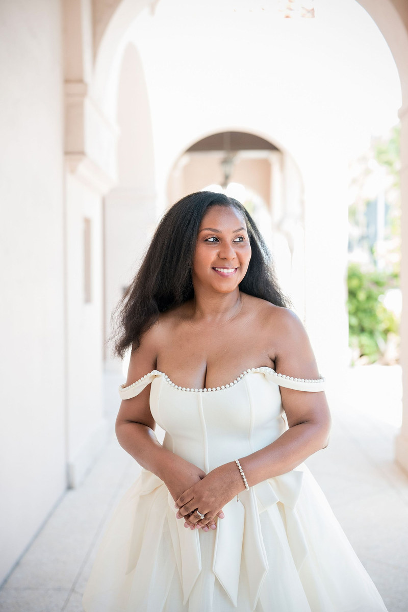 Woman in a white off-the-shoulder dress smiling in a sunlit arched hallway